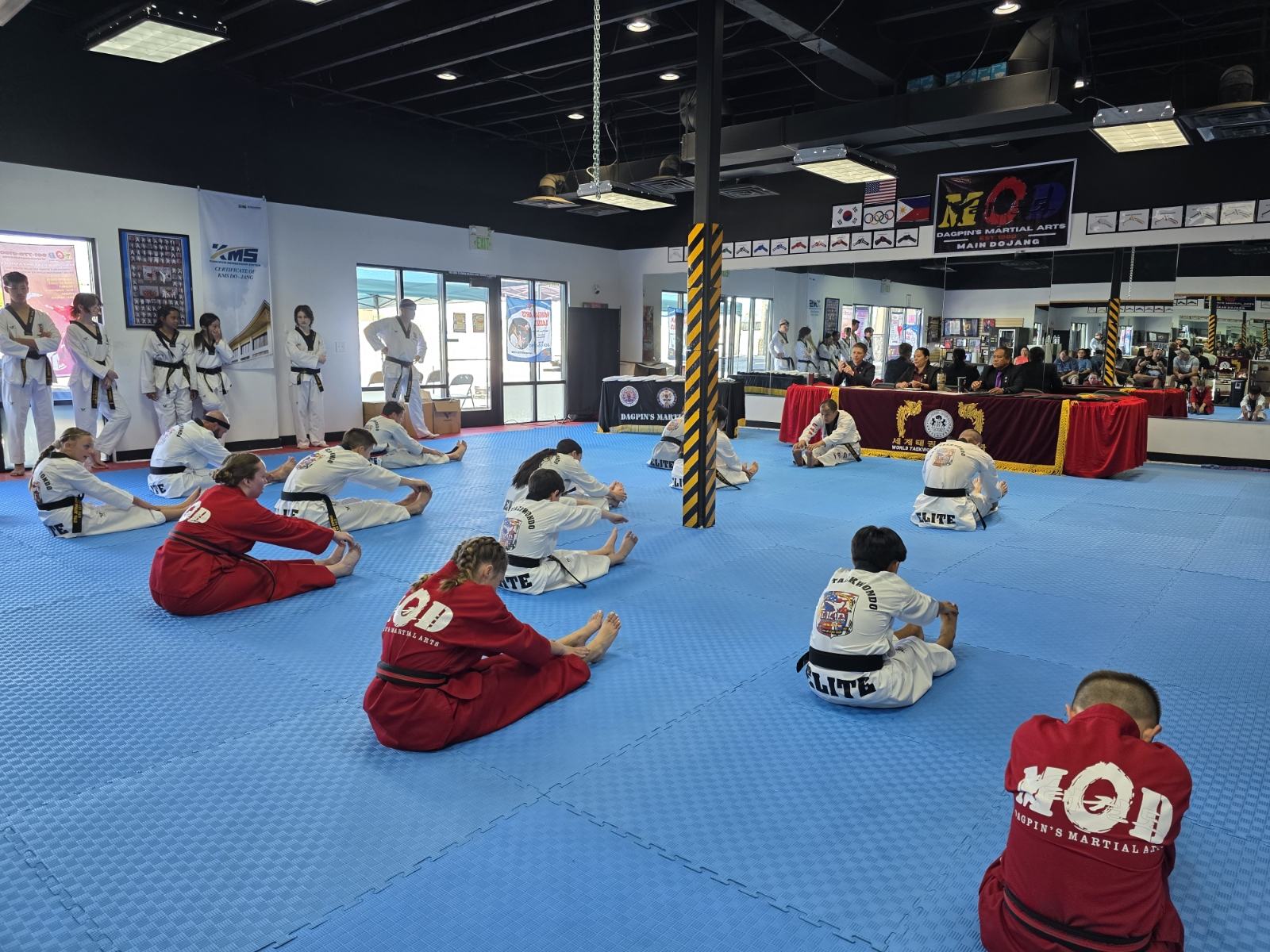 Students stretching on the mats before a belt testing event at MOD Taekwondo South Ogden, with judges seated at the head table