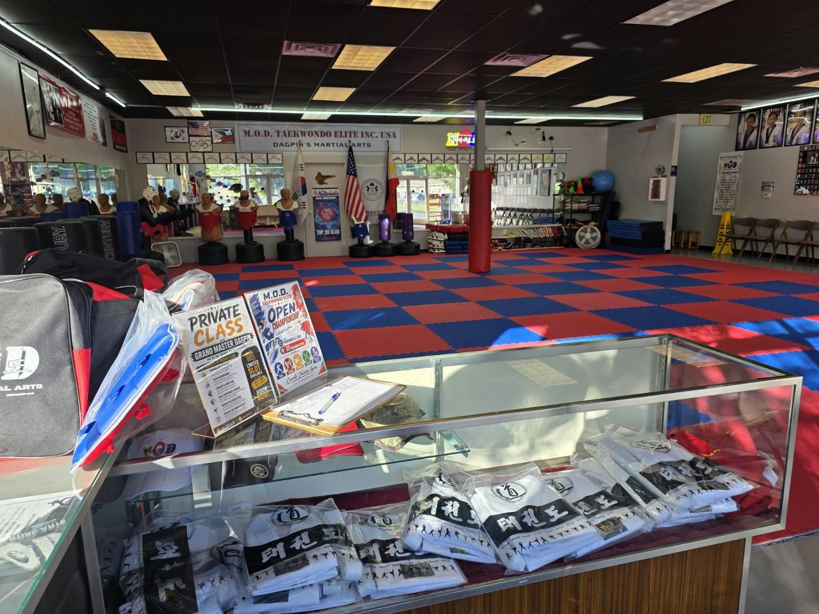 View across the MOD Taekwondo Harrisville training floor and pro-shop counter, with mats, bags and uniforms on display